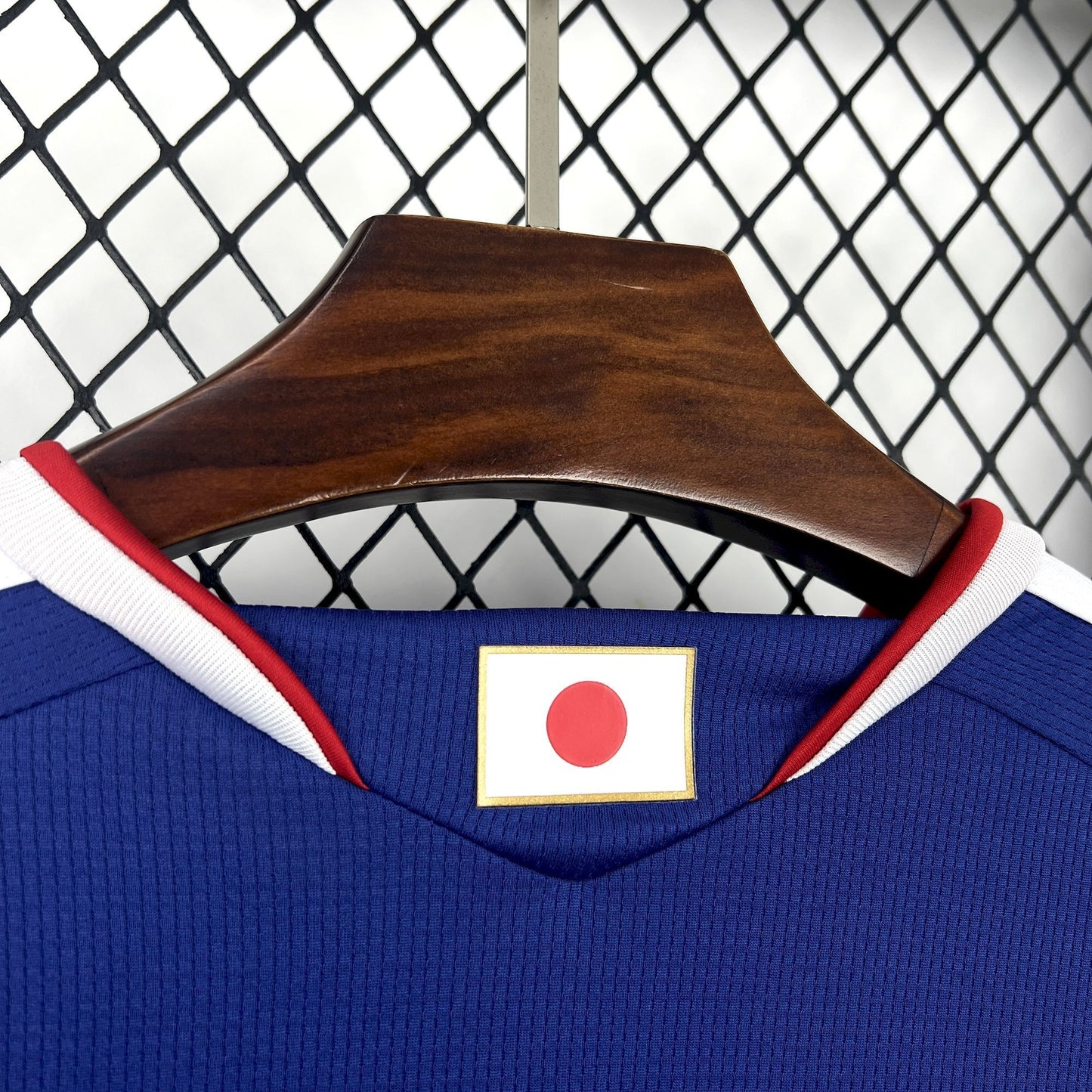 Japan 2026 Home Blue Jersey - Blue sports jersey with Japanese flag patch on a wooden hanger against a chain-link fence background.