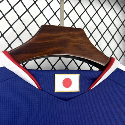 Japan 2026 Home Blue Jersey - Blue sports jersey with Japanese flag patch on a wooden hanger against a chain-link fence background.