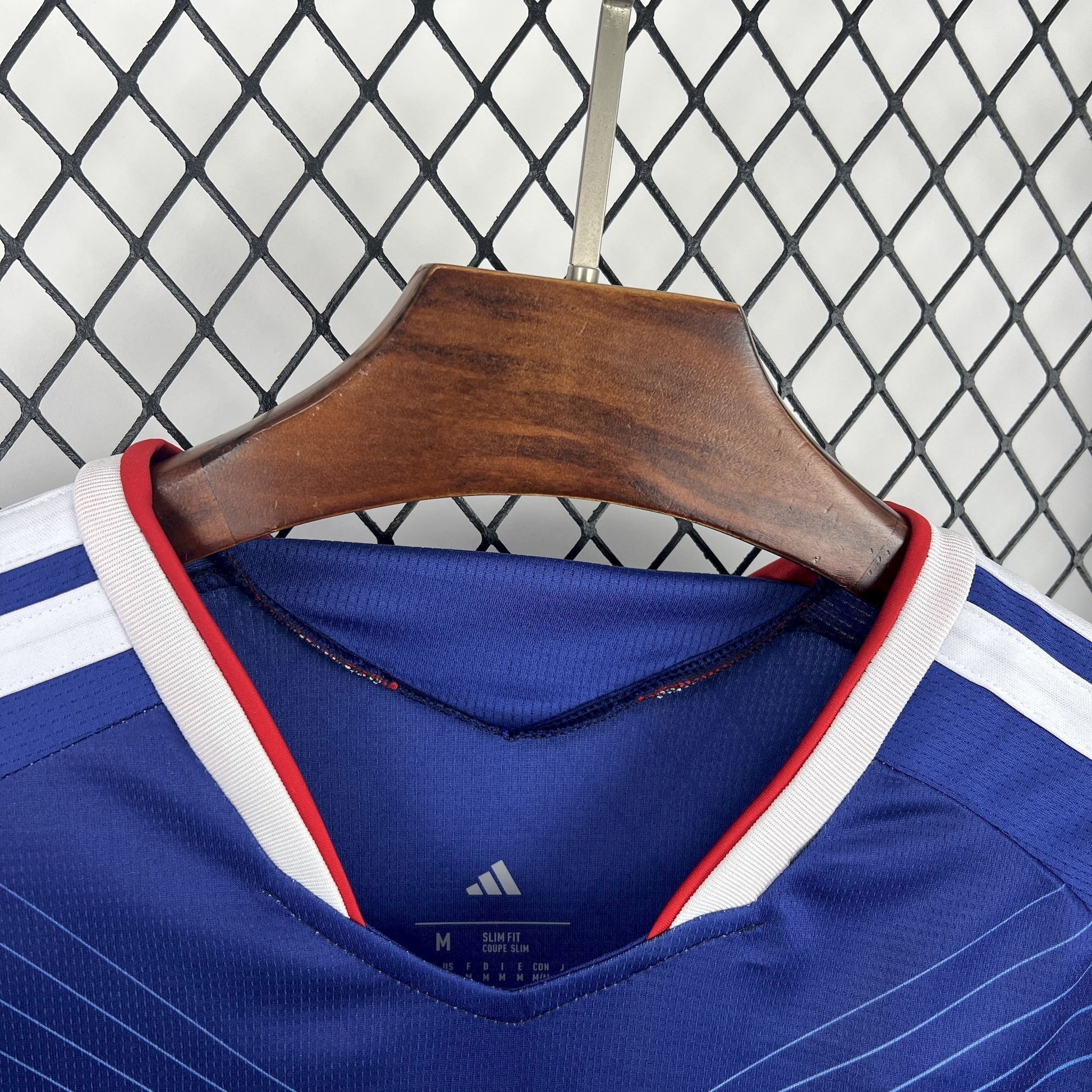 Japan 2026 Home Blue Jersey - Blue sports jersey with red and white accents on a wooden hanger against a chain-link fence background.